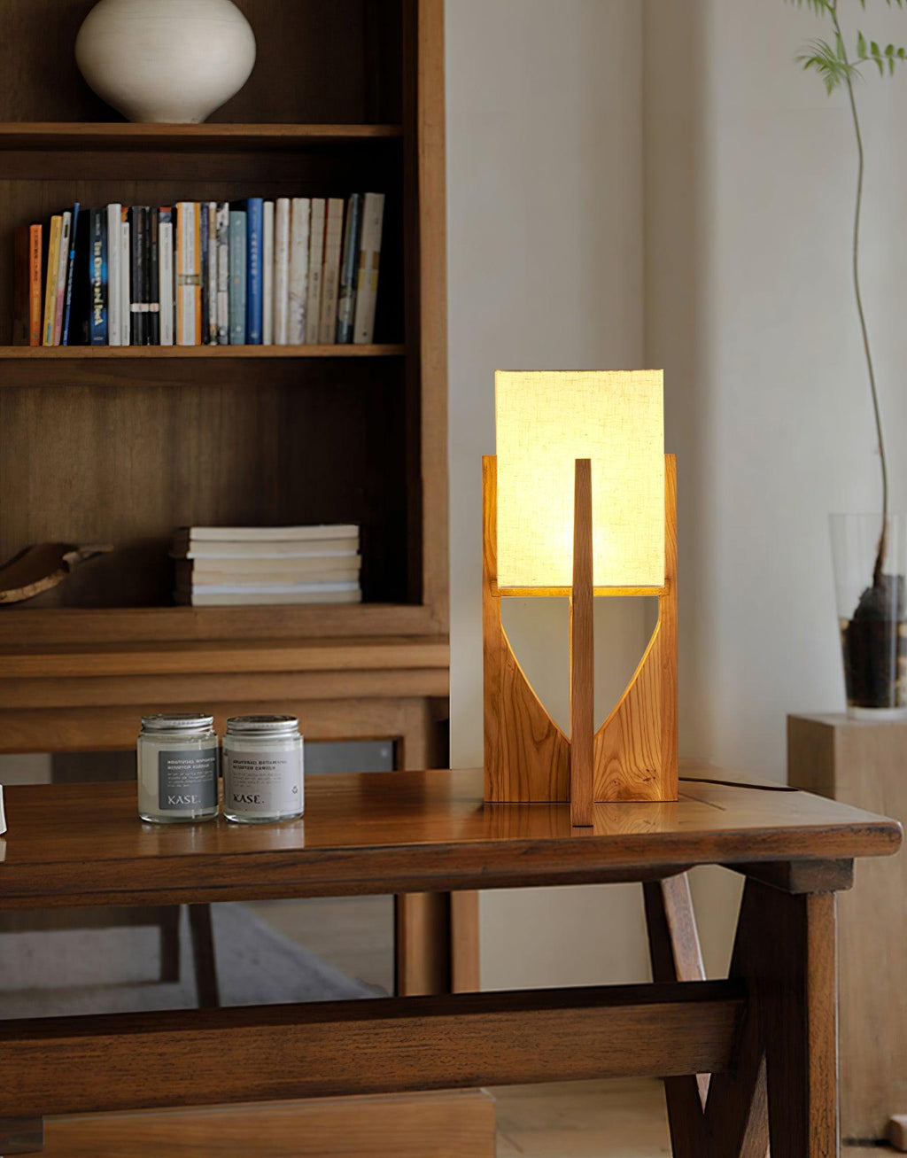 Small Clérachic table lamp lit on a wooden console, with a bookshelf, two jars and a tall branch in the background.