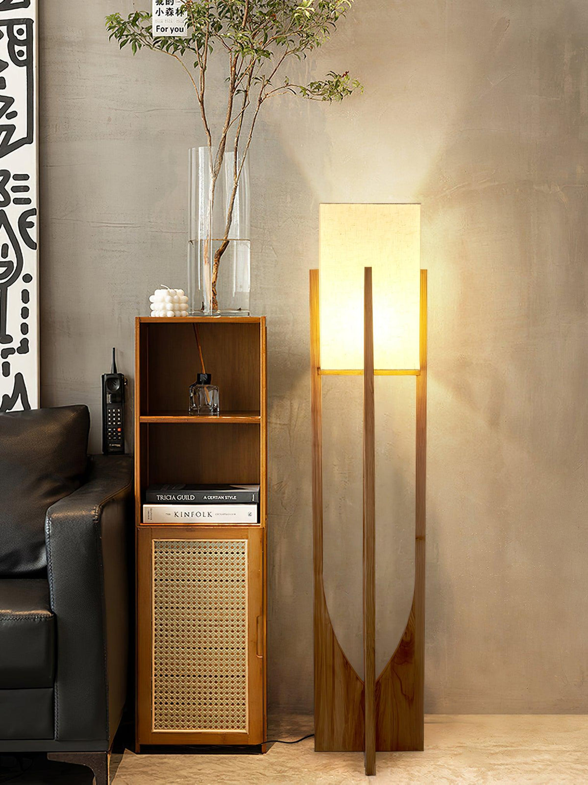 Tall Clérachic floor lamp glowing beside a narrow cane-front cabinet with a glass vase of branches, near a black leather sofa.