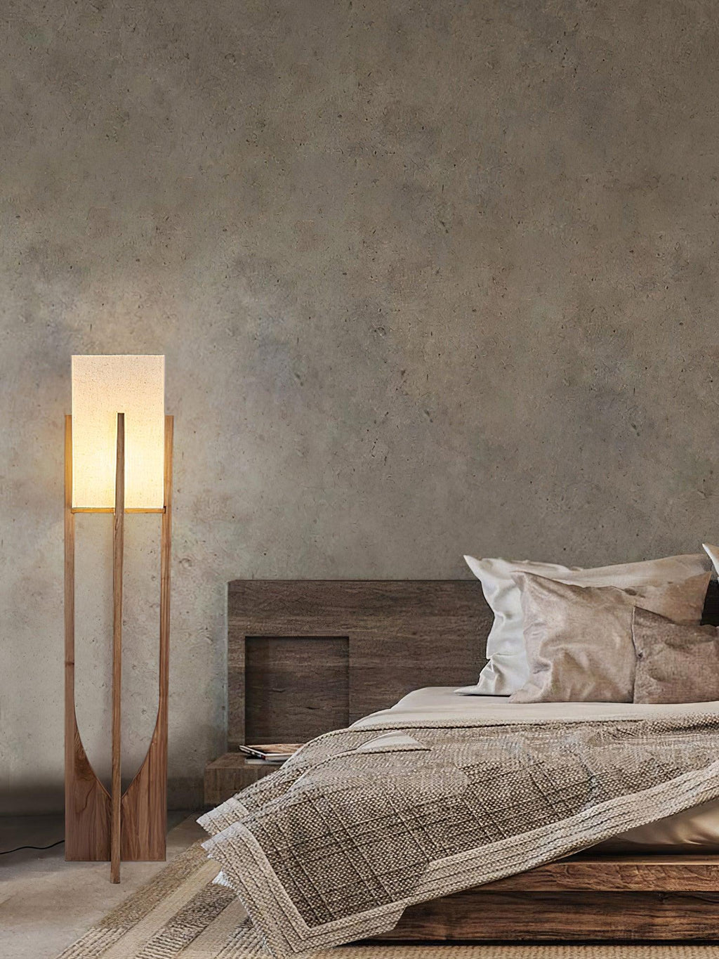 A Clérachic floor lamp stands beside a low wooden bed with rumpled linen, against a textured grey plaster wall.