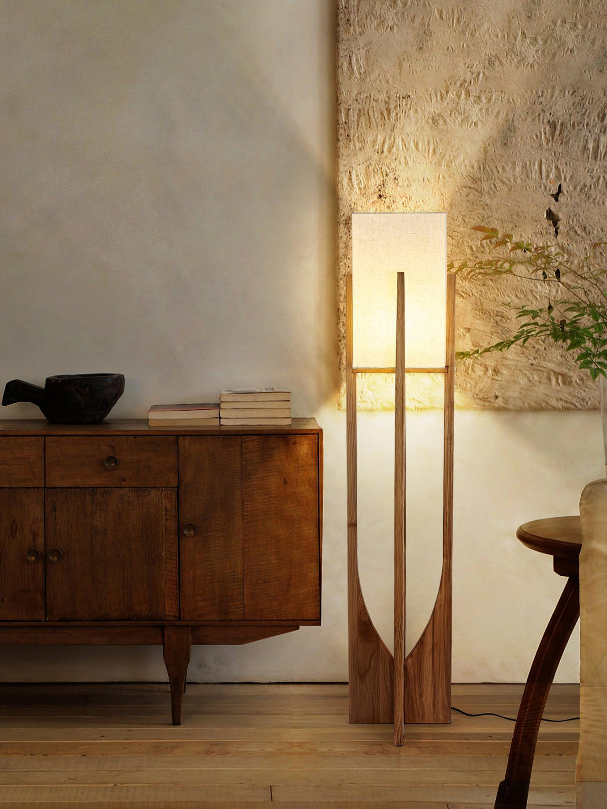 Lit Clérachic floor lamp beside a wooden cabinet with a dark bowl, with a rough plaster wall glowing above it.
