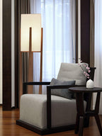 Tall Clérachic floor lamp, lit, beside a grey armchair and a round side table, in front of sheer curtains.