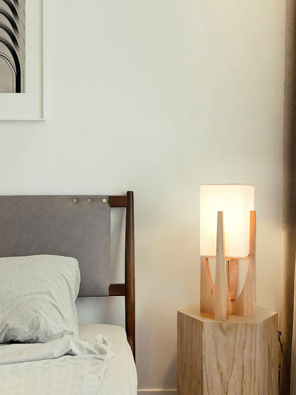 Small Clérachic table lamp in natural wood, lit, on a pale wooden block beside a bed with grey bedding.