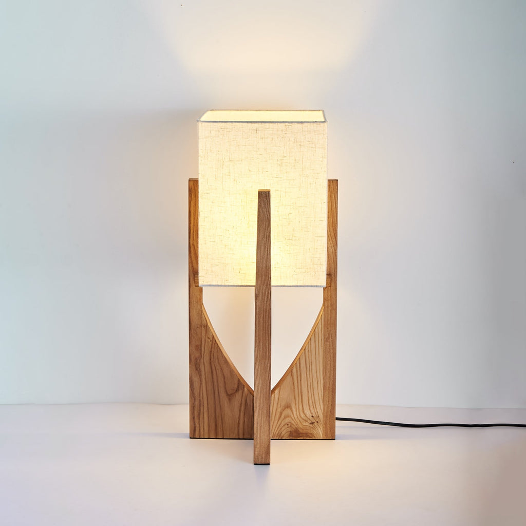 Short Clérachic table lamp in natural wood, with a lit fabric shade and a black cord, on a pale backdrop with a warm glow above.