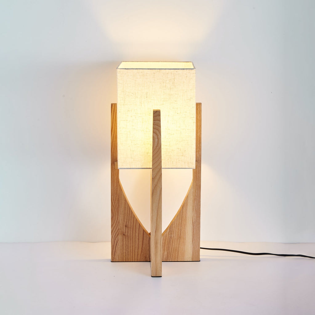 Short natural wood Clérachic lamp, lit, with a warm glow on the wall behind and a black cord trailing on the floor.