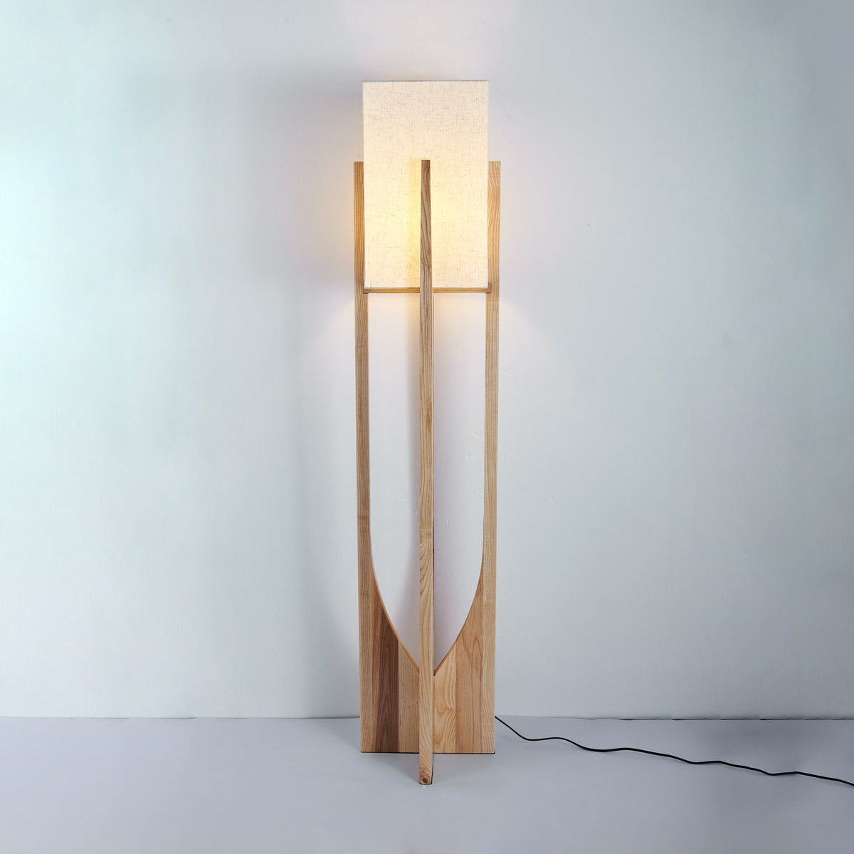 Tall Clérachic floor lamp in light natural wood, its shade unlit, against a pale blue wall with a black cord on the floor.
