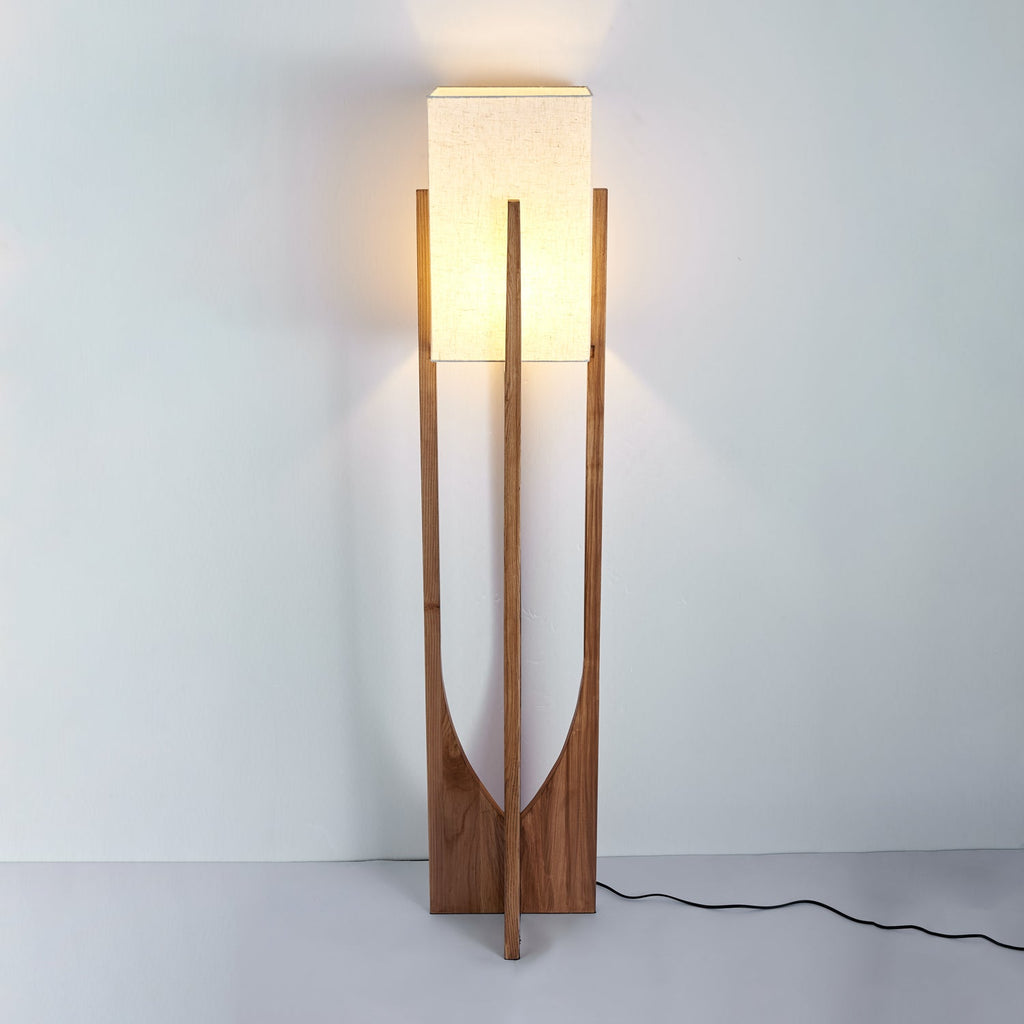 Tall Clérachic floor lamp in a darker wood, lit warmly, with a glow on the wall above and a black cord on the floor.