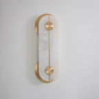 Clérachic Auriel wall light seen straight on, its alabaster capsule cradled by two brushed brass crescents on a pale grey wall.