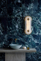 The Clérachic sconce lighting a bathroom of dark green marble, mounted above a stone basin and a brass wall tap.