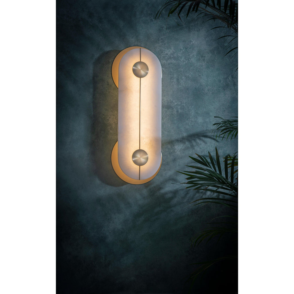 The Clérachic wall light glowing on a deep teal wall beside palm fronds, alabaster diffusing the light into a soft halo.