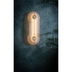 The Clérachic wall light glowing on a deep teal wall beside palm fronds, alabaster diffusing the light into a soft halo.