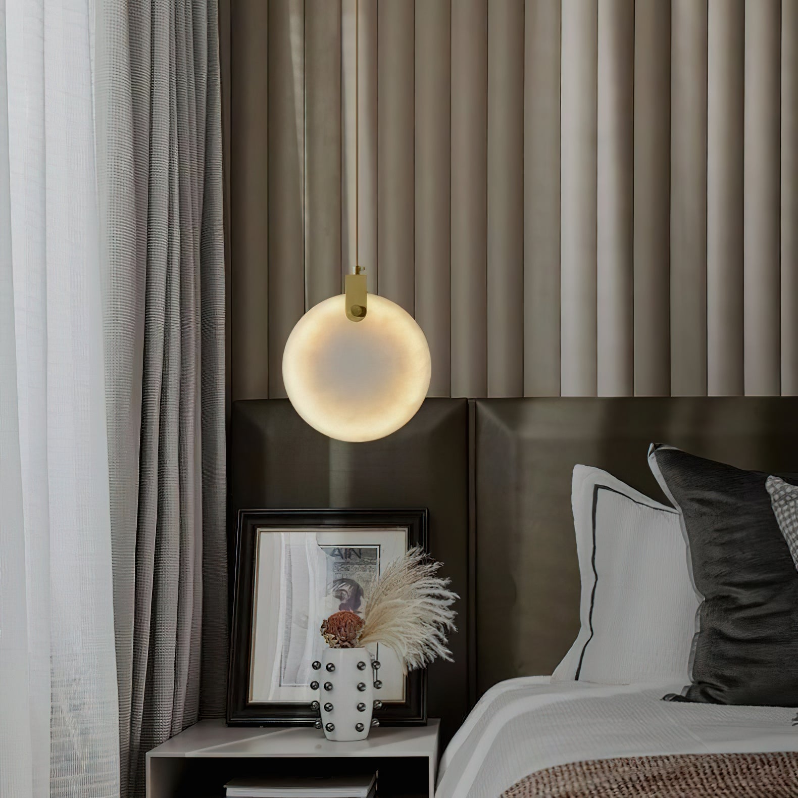 The Clérachic alabaster pendant beside a bed, its lit disc reading as a small moon against a ribbed headboard wall.