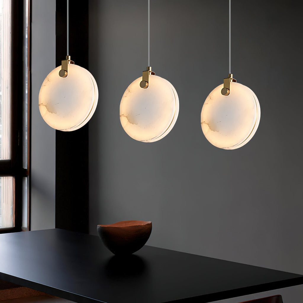 Three Clérachic alabaster pendants in a row above a black dining table, each disc lit from between its two stone faces.