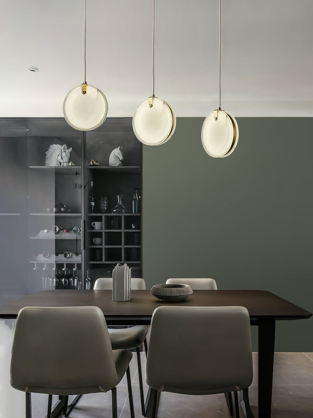 Three Clérachic alabaster pendants over a dark dining table, hung at staggered drops against a deep green wall.