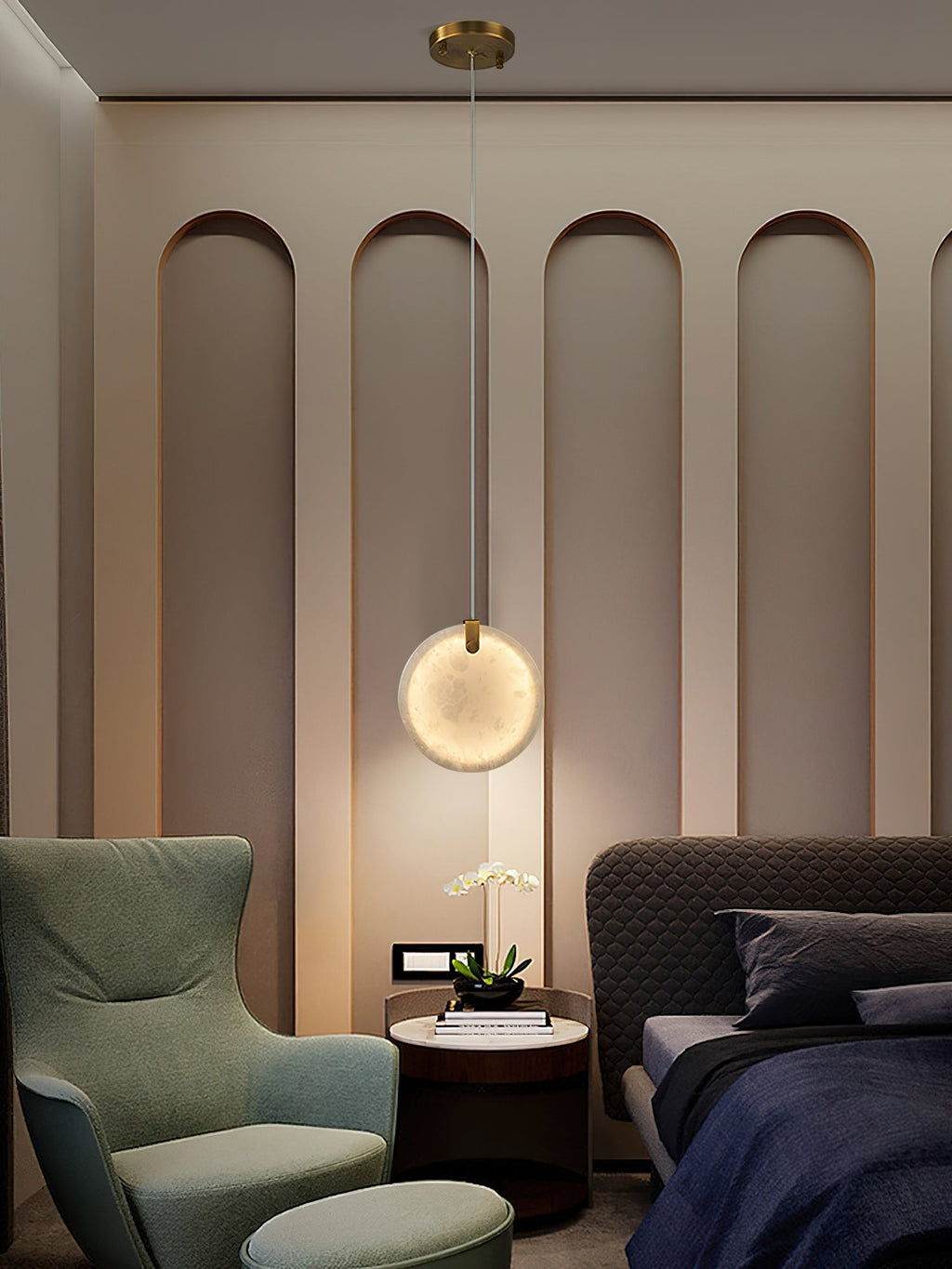 A Clérachic alabaster disc pendant lit low over a round bedside table, between a green wing chair and a quilted bed.