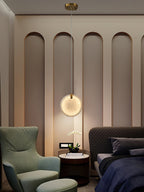 A Clérachic alabaster disc pendant lit low over a round bedside table, between a green wing chair and a quilted bed.