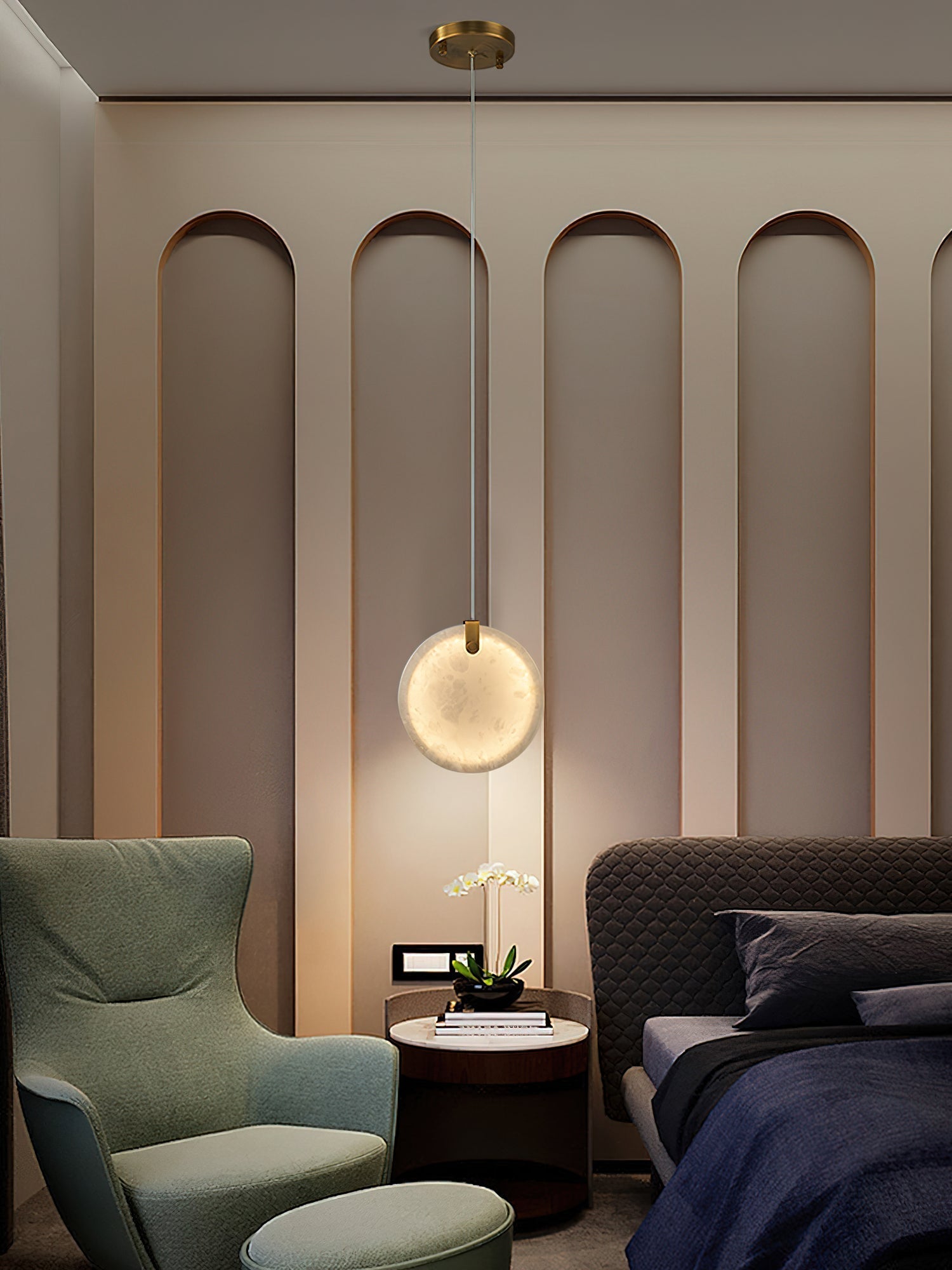 A Clérachic alabaster disc pendant lit low over a round bedside table, between a green wing chair and a quilted bed.