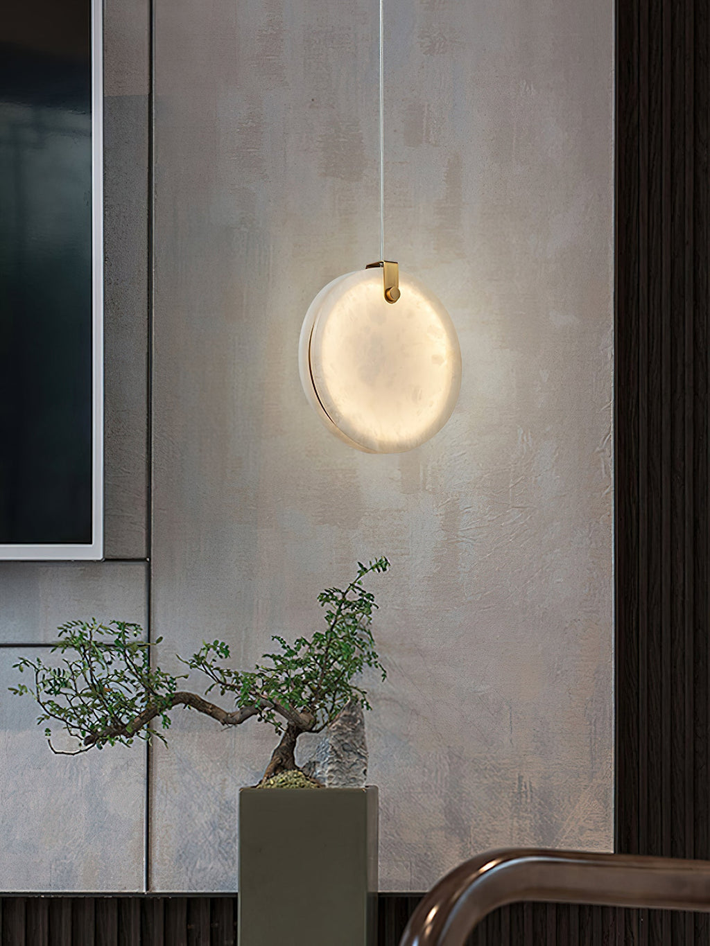 A single Clérachic alabaster pendant lit against a textured plaster wall, throwing a soft halo above a bonsai.