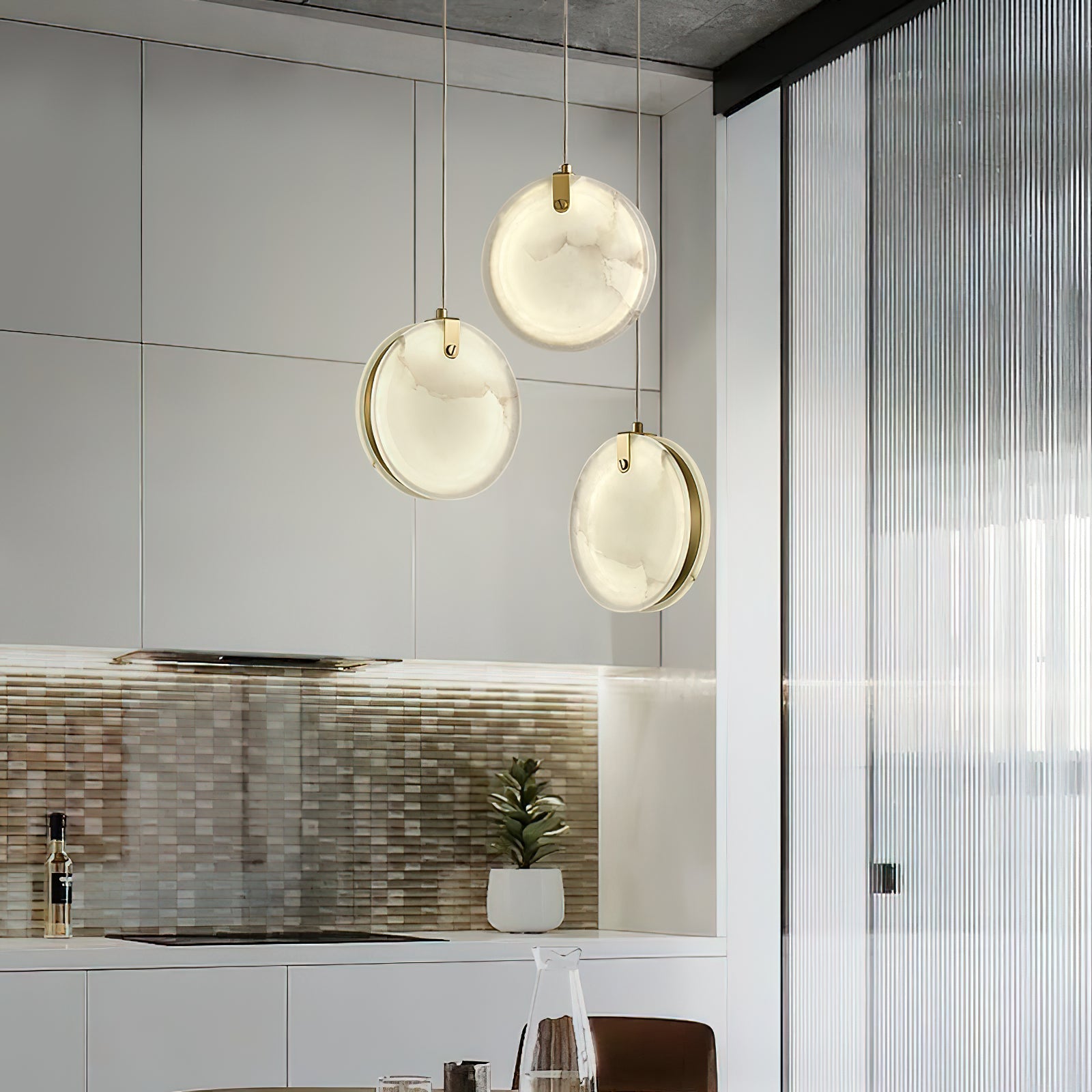 Three Clérachic alabaster pendants clustered in a bright kitchen, brass rims visible behind each glowing disc.