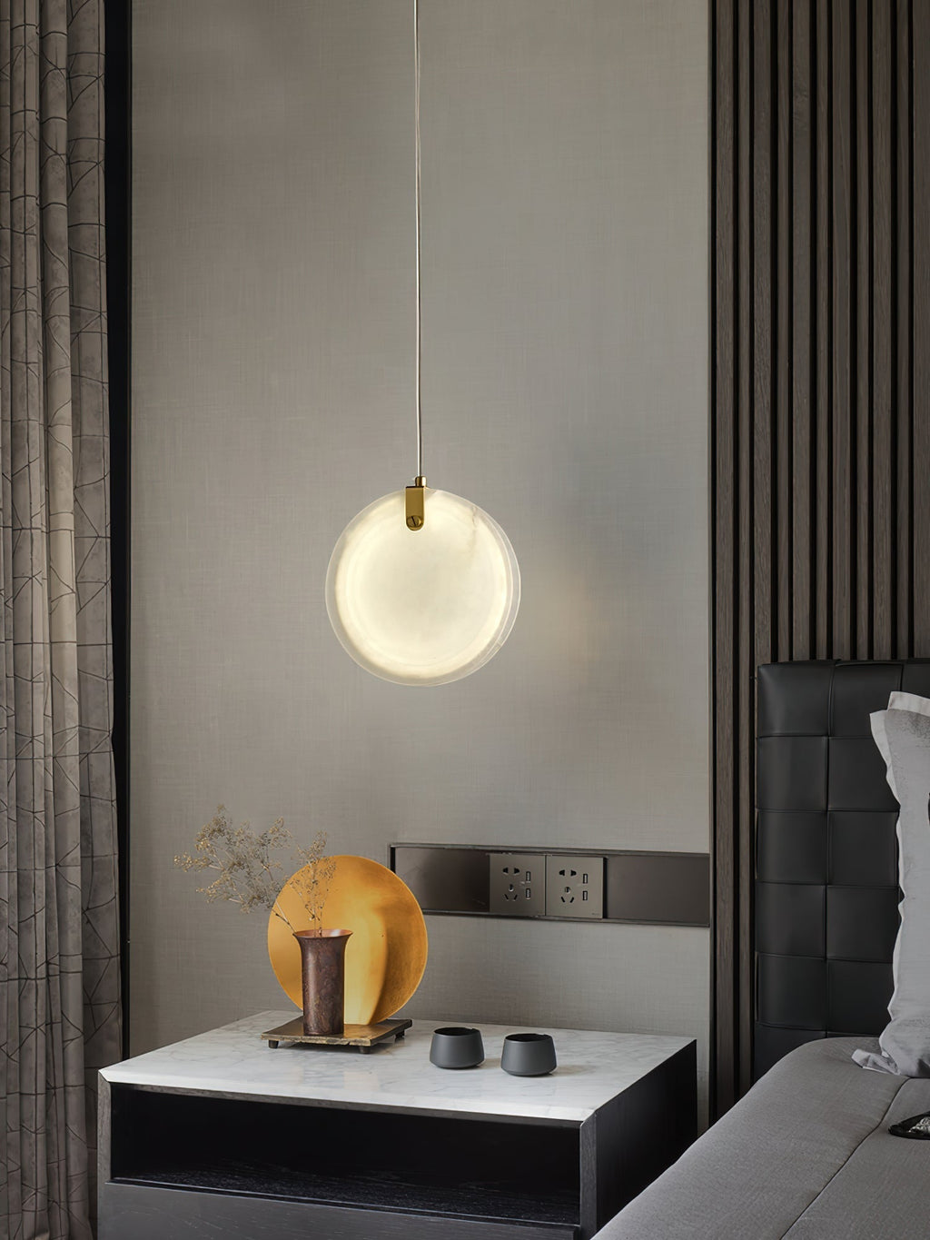 A Clérachic alabaster pendant lit over a marble bedside table, the brass strap catching light at the top of the disc.