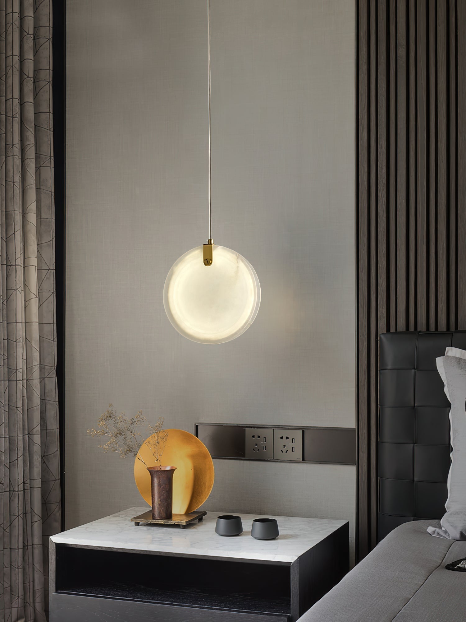 A Clérachic alabaster pendant lit over a marble bedside table, the brass strap catching light at the top of the disc.