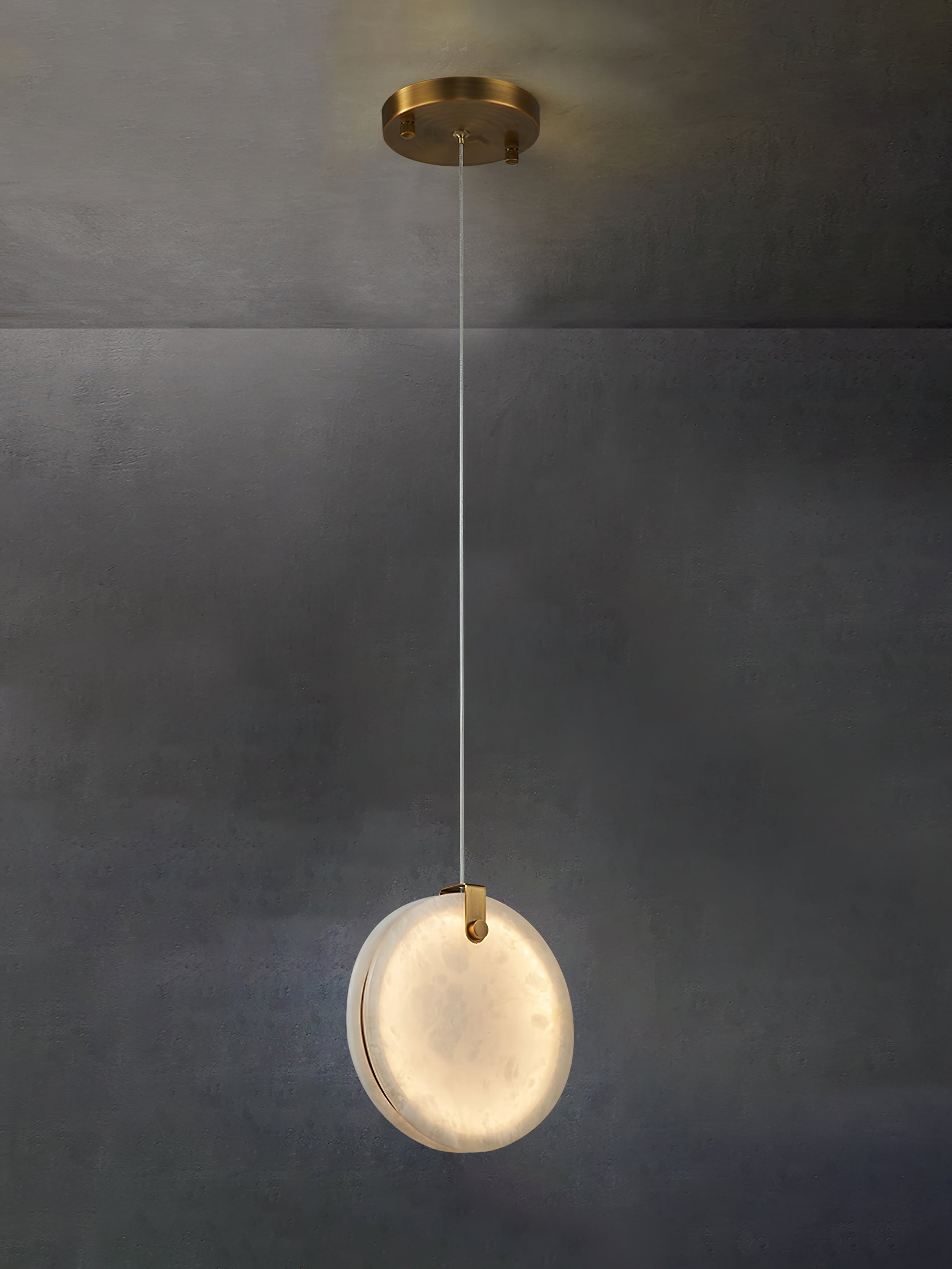 A Clérachic alabaster pendant lit on its long cable, the stone warming to ivory and honey against a concrete wall.