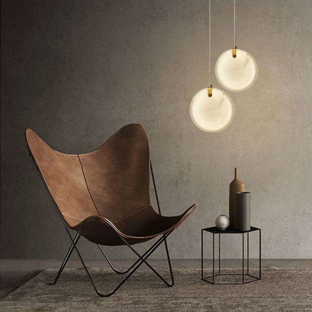 Two Clérachic alabaster pendants lit beside a brown leather butterfly chair against a raw concrete wall.