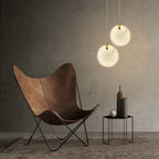 Two Clérachic alabaster pendants lit beside a brown leather butterfly chair against a raw concrete wall.