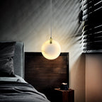 A Clérachic alabaster pendant lit at a bedside, blind shadows striping the wall behind the glowing disc.