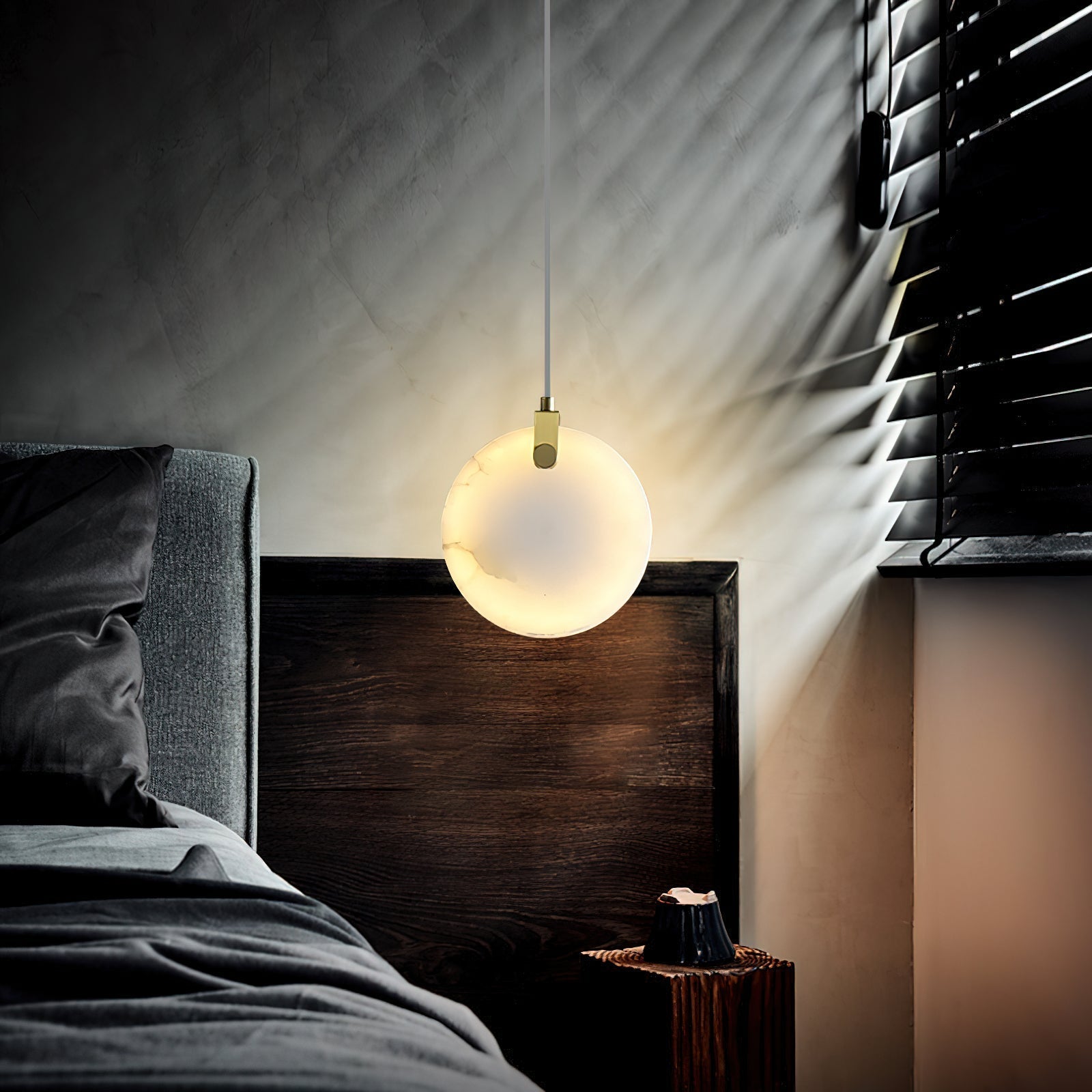A Clérachic alabaster pendant lit at a bedside, blind shadows striping the wall behind the glowing disc.