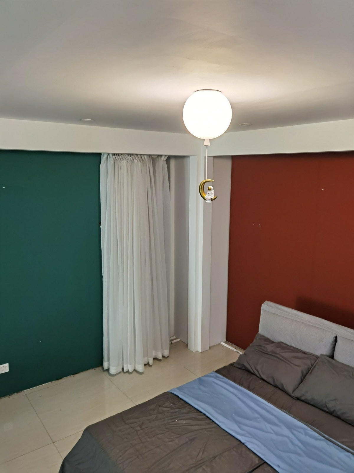 One white Clérachic balloon light with a small moon-shaped ornament, above a bed between green and rust-red walls.