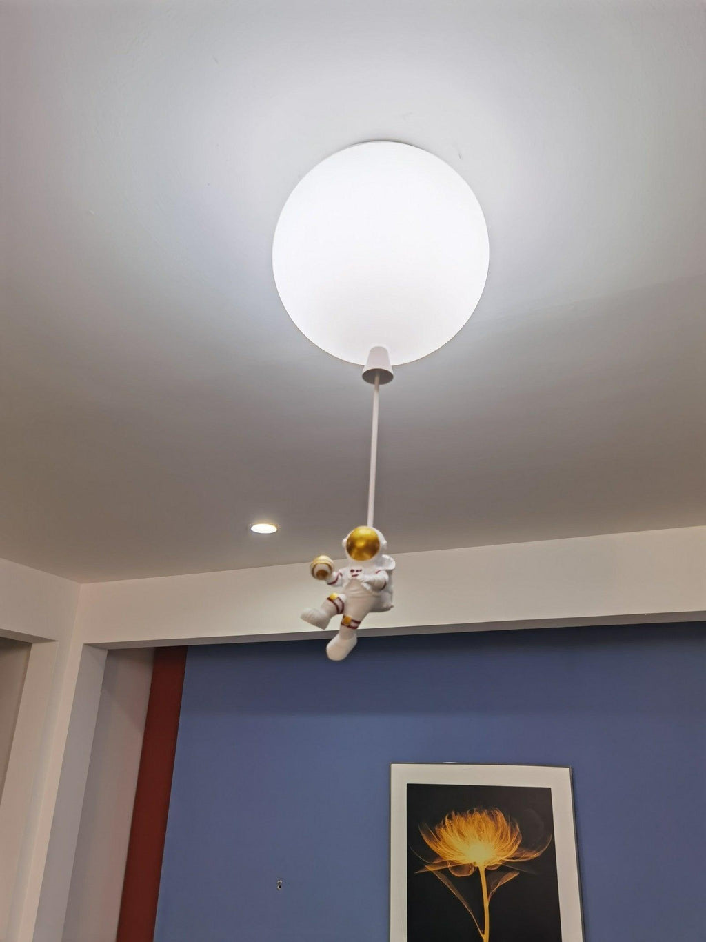 Single white Clérachic balloon light on a long cord, its astronaut perched low, against a ceiling with downlights and a blue wall.