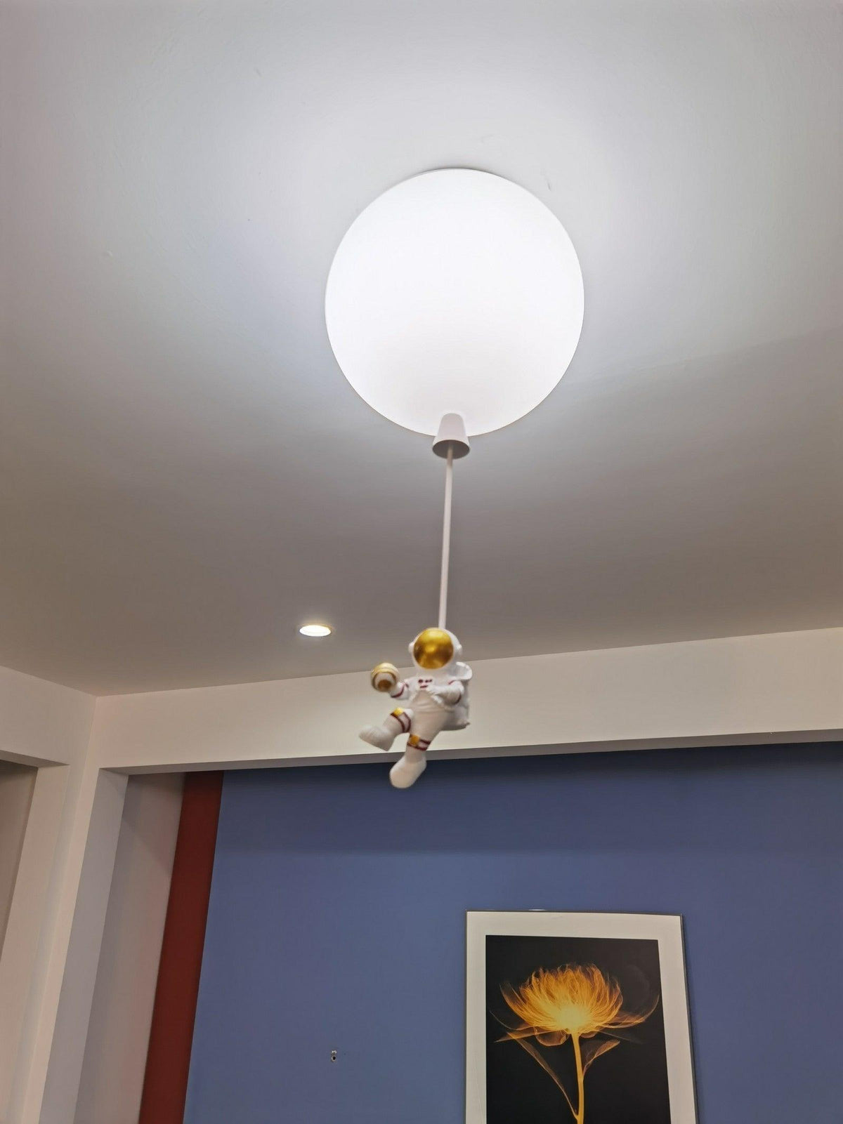 Single white Clérachic balloon light on a long cord, its astronaut perched low, against a ceiling with downlights and a blue wall.