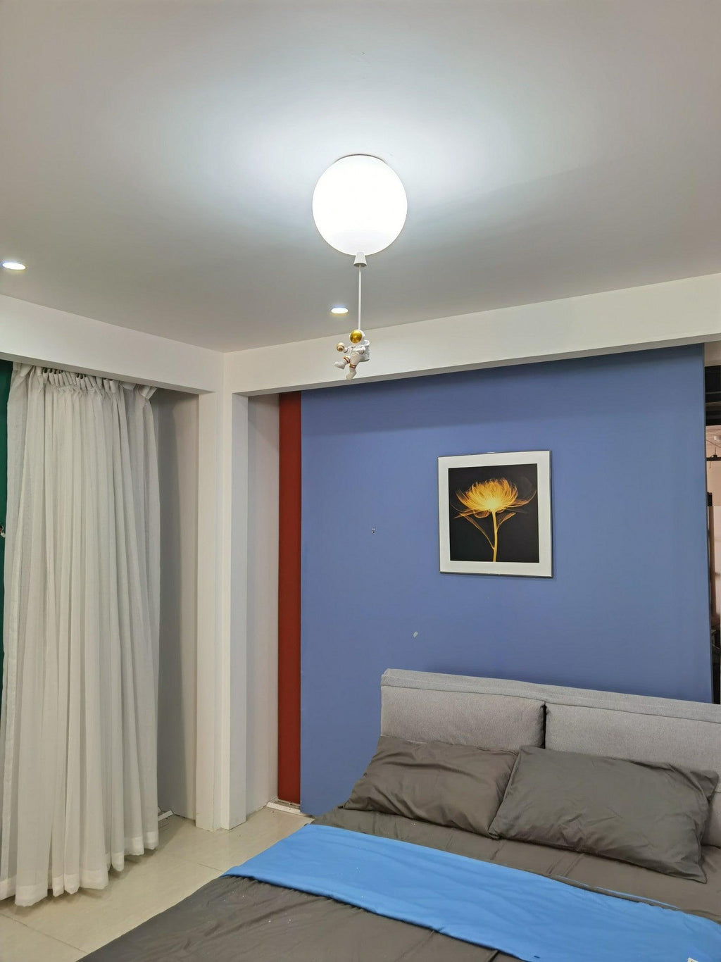 One white Clérachic balloon ceiling light above a bed with a blue throw, a blue feature wall and a framed flower print.