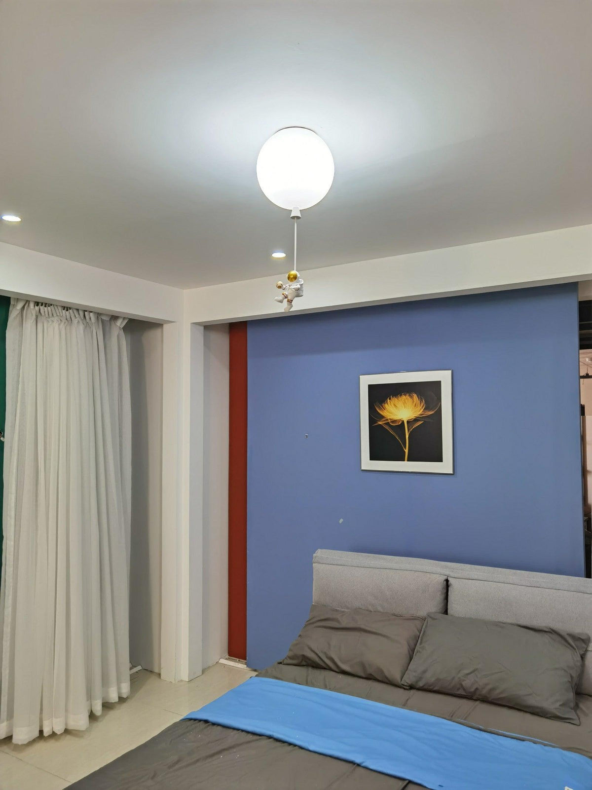 One white Clérachic balloon ceiling light above a bed with a blue throw, a blue feature wall and a framed flower print.