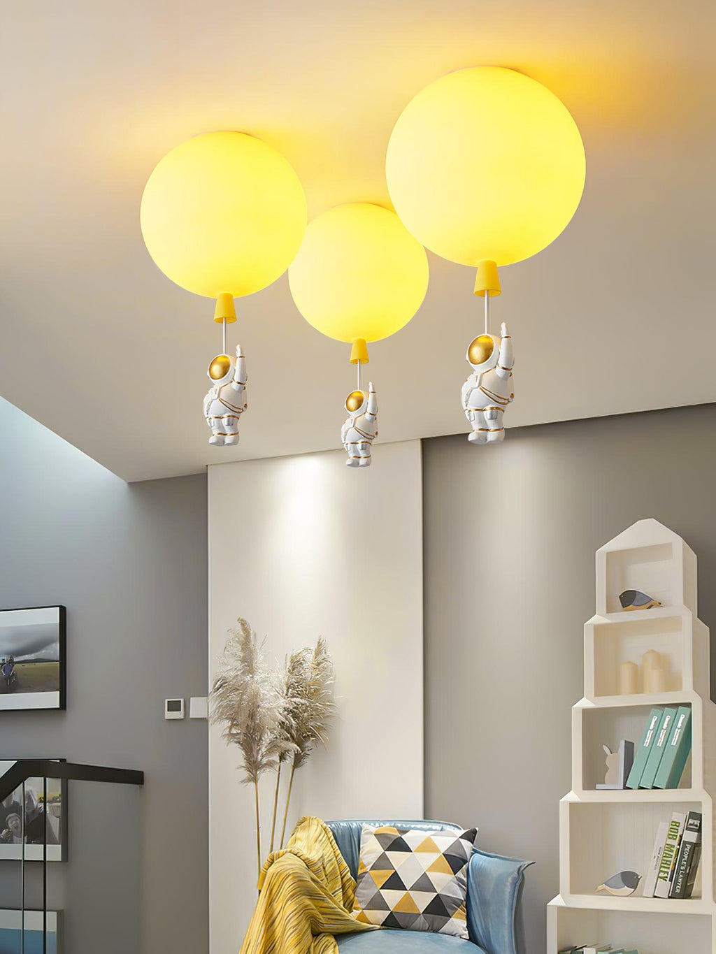 Three yellow Clérachic balloon lights with astronauts in a room with a white house-shaped shelf and a blue chair.