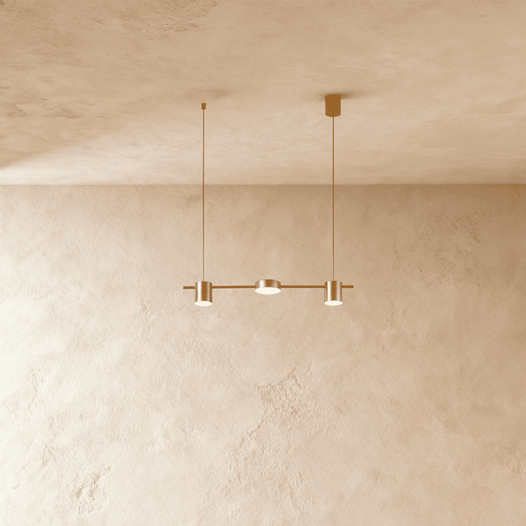 Angled view of the Clérachic gold three-head pendant bar, cylindrical heads glowing warm white beneath a textured ceiling.