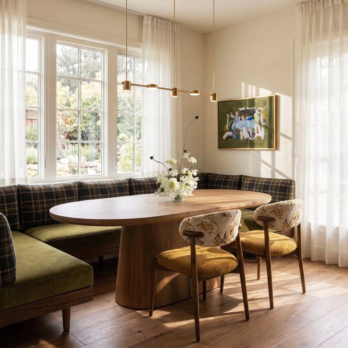 The Clérachic gold five-head pendant bar glowing warm white above a plaid banquette and oval dining table by tall windows.