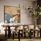An unlit Clérachic gold bar pendant light hangs above a long dining table beneath a framed landscape painting.