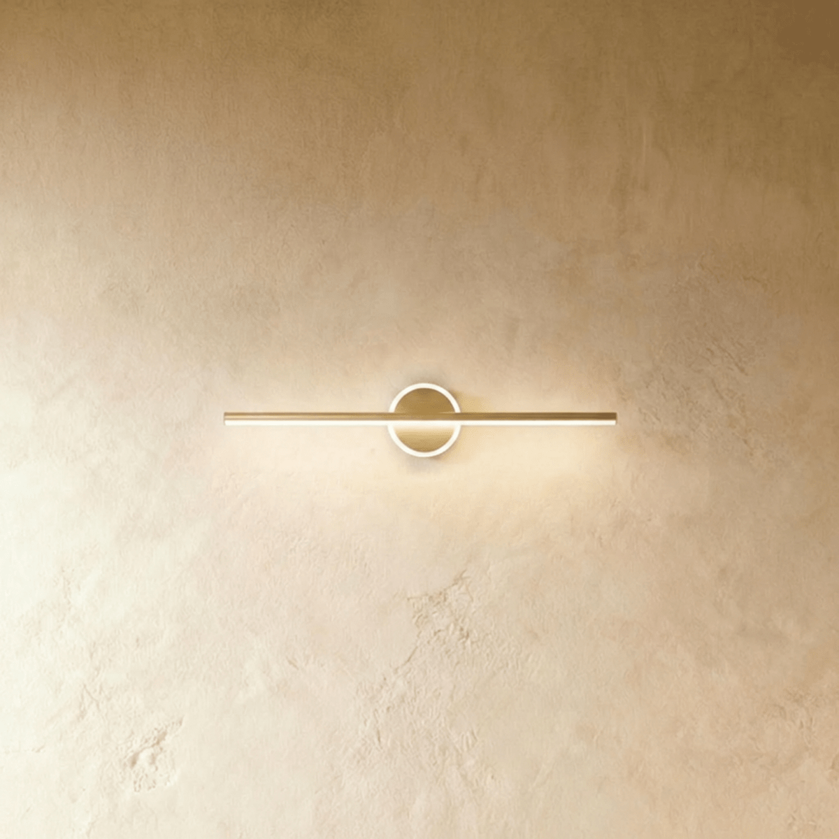 Clérachic slim gold bar wall light crossing a round backplate with a softly glowing ring on a limewash wall.