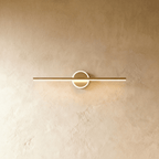 Clérachic longer gold bar wall light with a bright ring around its backplate, on a warm textured wall.