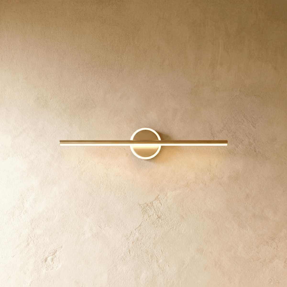 Clérachic longer gold bar wall light with a bright ring around its backplate, on a warm textured wall.