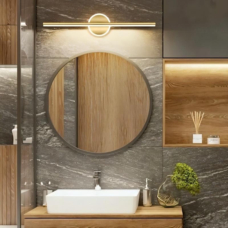 Clérachic gold bar wall light above a round bathroom mirror and white basin, with grey stone tiles and warm wood.