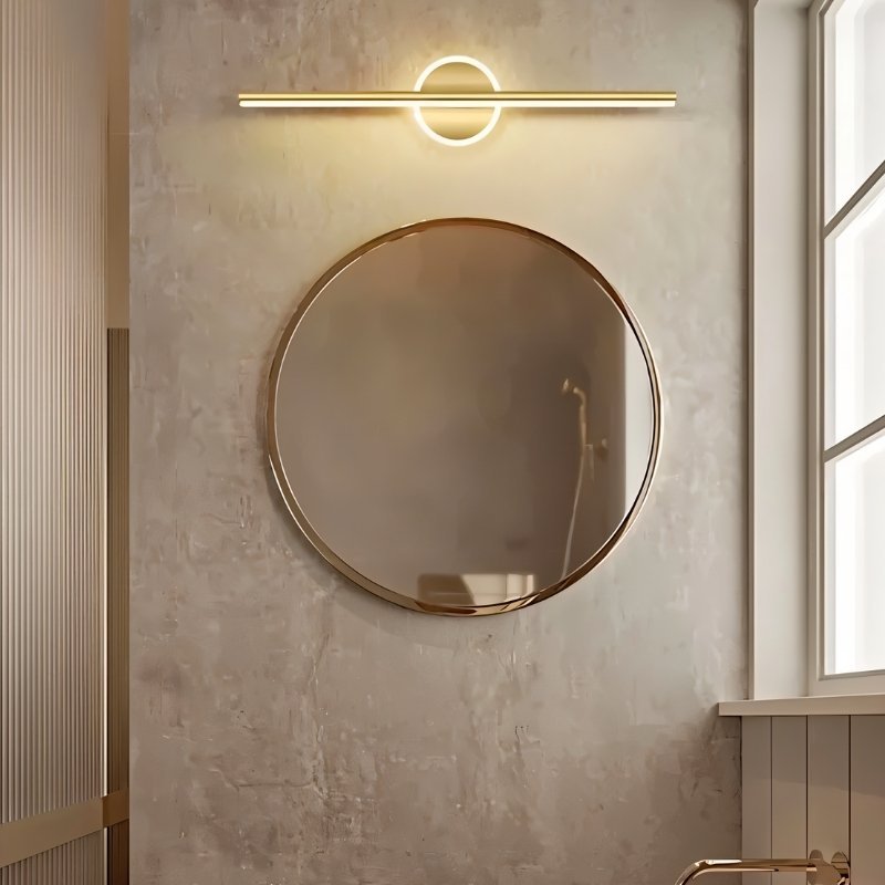 Clérachic gold bar wall light above a round gold-framed mirror, on a textured concrete-look wall beside a window.