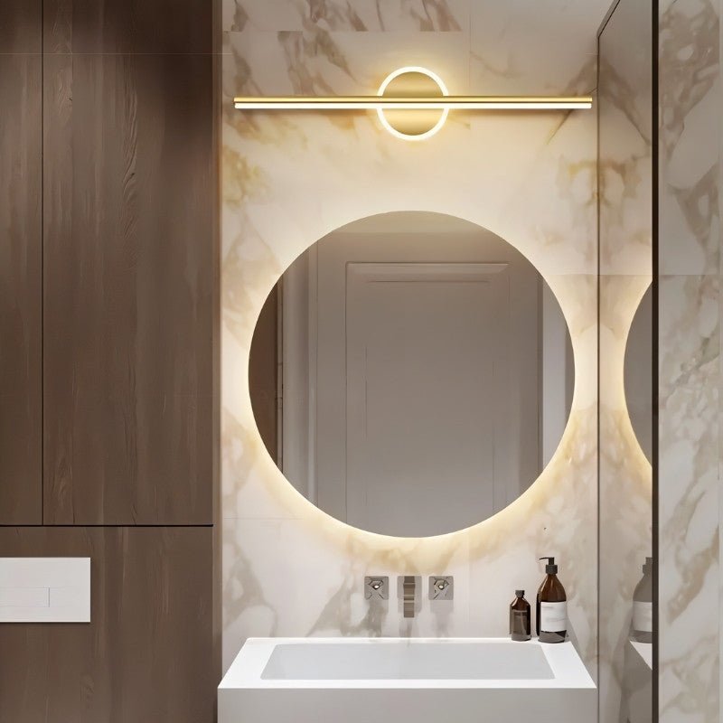 Clérachic gold bar wall light over a backlit round mirror in a marble-tiled bathroom with a white basin.