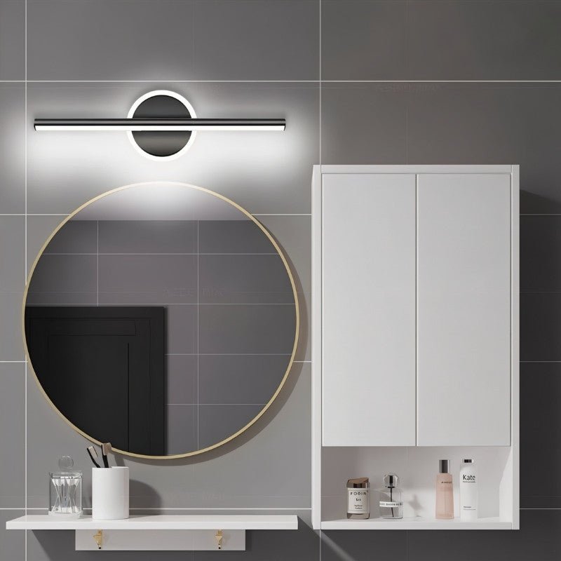 Clérachic black bar wall light lit cool white above a round gold-framed mirror, with a white cabinet in a bathroom.
