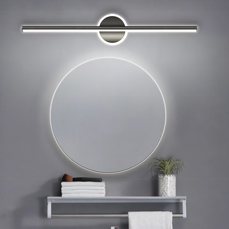 Clérachic black bar wall light above a round backlit mirror, a towel shelf and a small plant in a grey bathroom.
