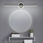 Clérachic black bar wall light above a round backlit mirror, a towel shelf and a small plant in a grey bathroom.