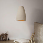 This Clérachic pendant light in a tapered bell shape hangs against a pale wall, above a wicker basket and a cushioned wooden chair.
