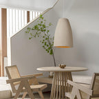 A beige Clérachic pendant light over a round wooden table with cane armchairs, beside a staircase and a leafy branch.