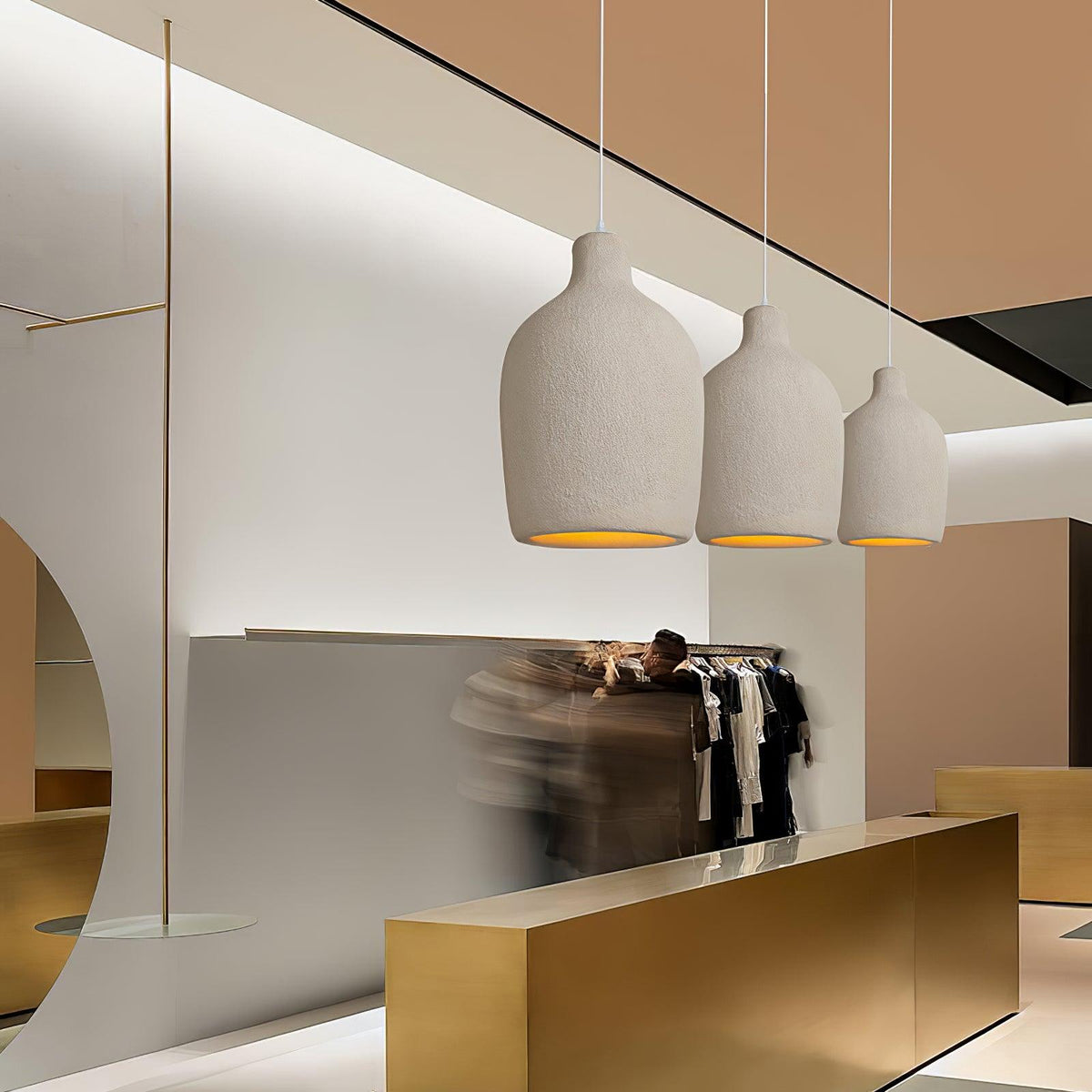 Three Clérachic pendant lights in graduated sizes hanging in a row above a gold counter in a bright clothes shop.